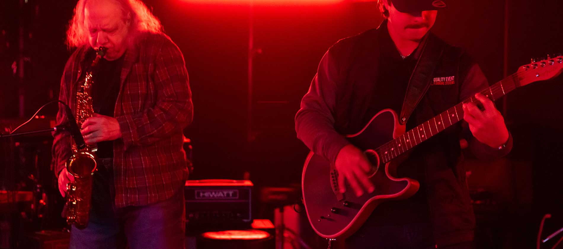 Saxophonist and guitarist playing side by side under red stage lights