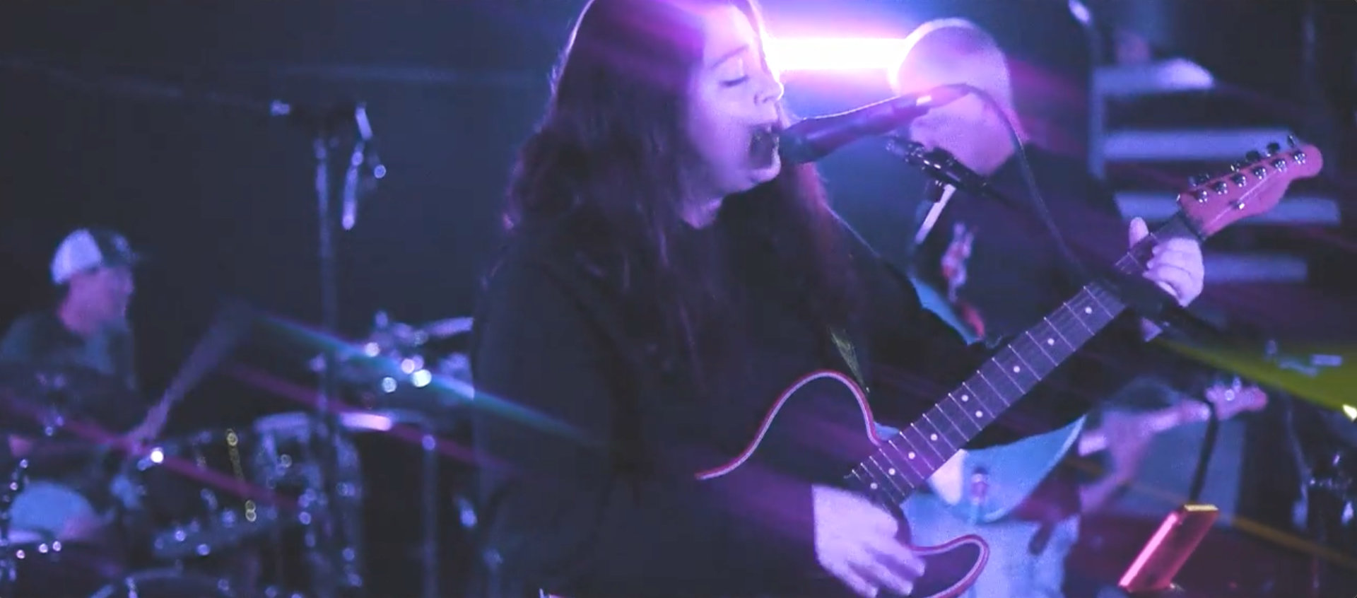 Singer with long hair singing and playing guitar under purple stage lights