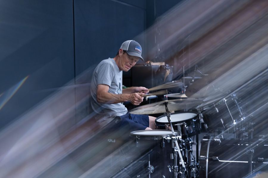 Drummer in a gray cap playing his kit, with light streaks rushing past