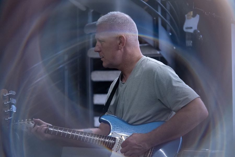 Guitarist playing a light-blue electric guitar, framed by a swirling lens effect