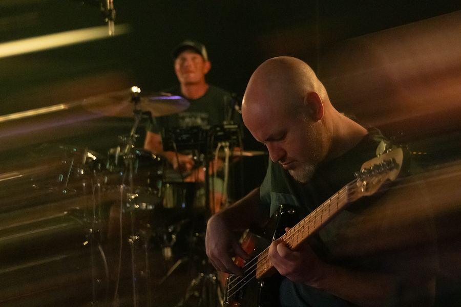 Bassist in the foreground with the drummer smiling behind him, light streaks across the frame