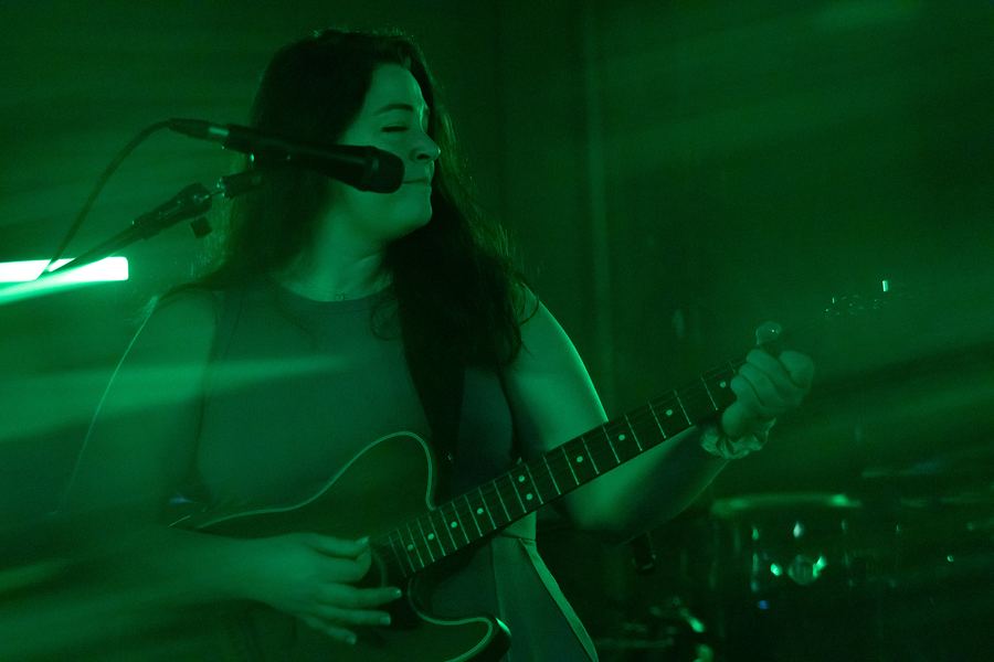 Singer with long dark hair singing and playing electric guitar under green light