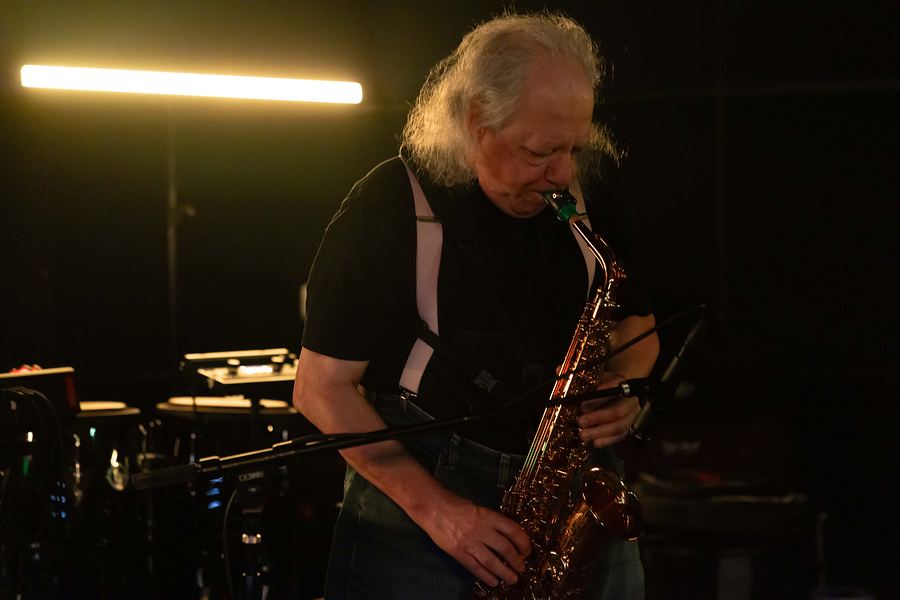 Saxophonist with long white hair playing alto sax under a warm light bar