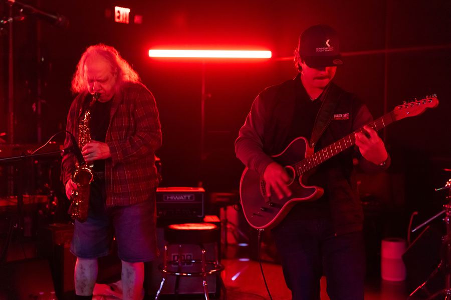 Saxophonist and guitarist playing side by side under red stage lights
