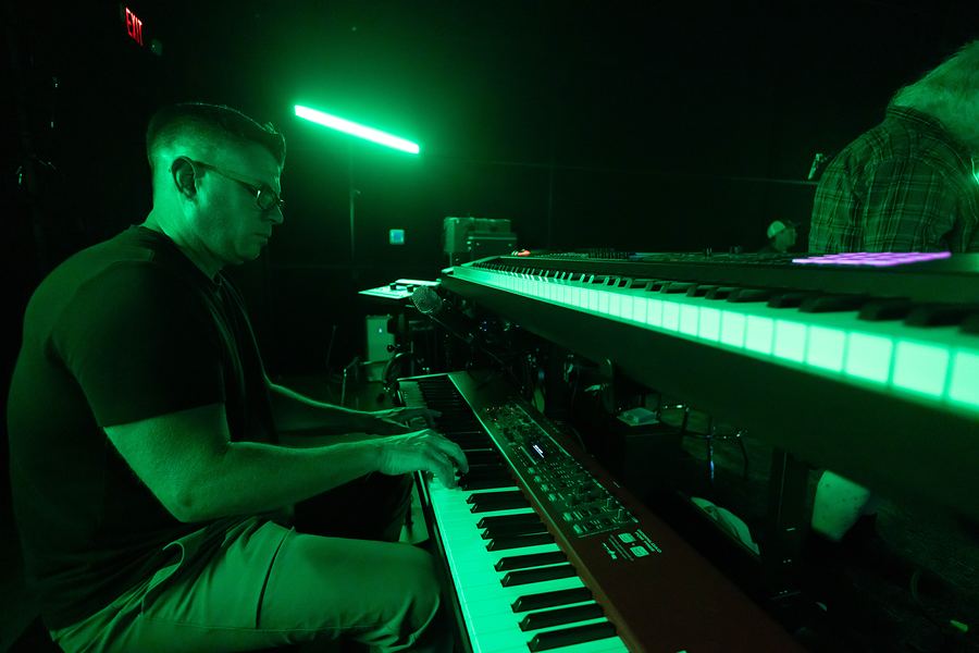 Keyboardist in glasses playing a stack of keyboards under green light
