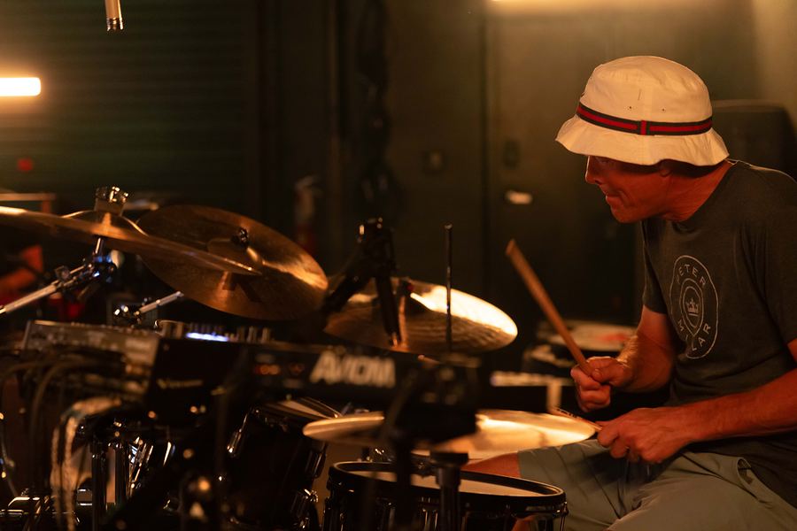 Smiling drummer in a bucket hat playing his kit in warm light