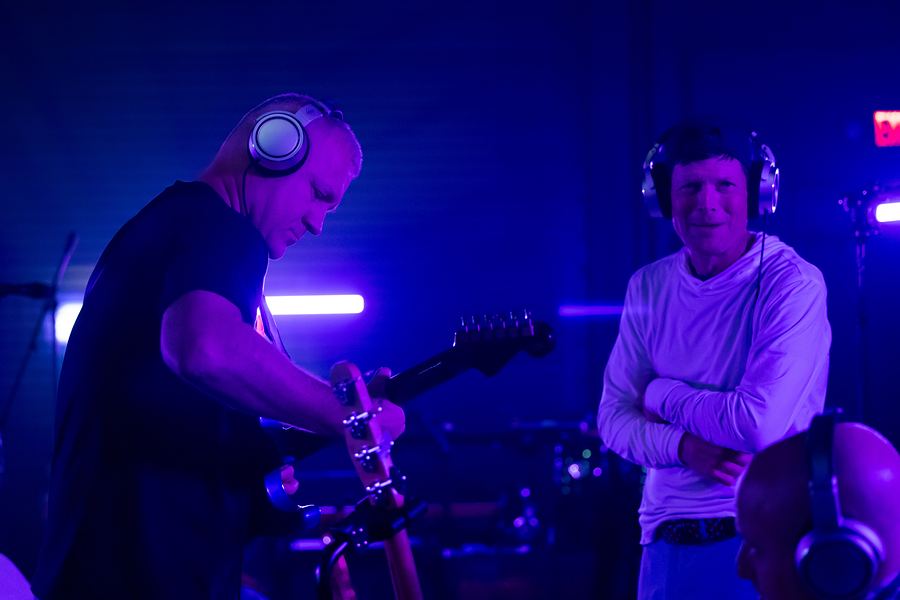 Guitarist in headphones beside a grinning bandmate with arms crossed, purple light
